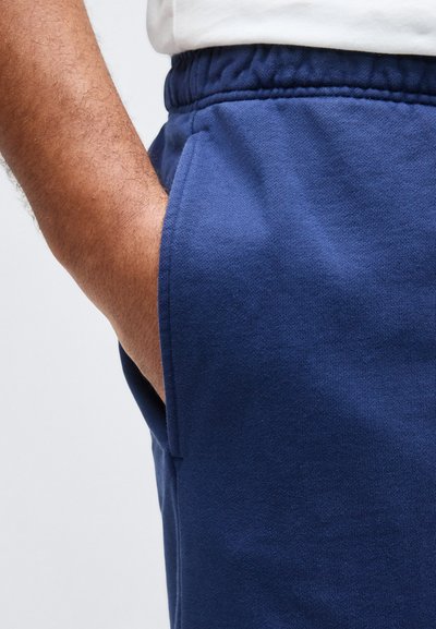 Ανδρικό χέρι που ακουμπά μέσα στην τσέπη μπλε σκούρων φόρμας με ελαστική μέση, φορεμένη με λευκό πουκάμισο.