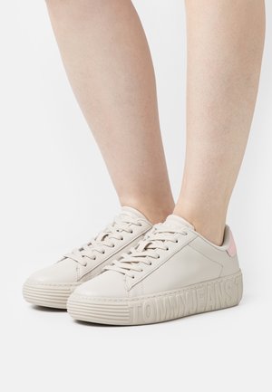 Béžové kožené tenisky s plochými tkaničkami, kulatou špičkou a texturovanou podrážkou. Na vnější straně je vyražené logo a na patě růžový akcent.