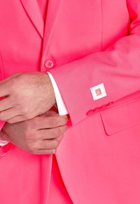 Giacca da abito rosa brillante con texture liscia, chiusura con un bottone, polsini di camicia bianca visibili e un'etichetta del marchio sulla manica.
