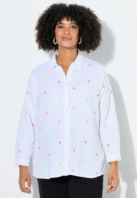 Frau mit lockigem Haar, die Perlen-Creolen trägt und ein weißes Hemd mit Knöpfen und Sternmustern in Orange, Pink und Grün, stehend vor einem weißen Hintergrund.