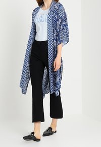 Kobieta w niebieskim, wzorowanym kimono z otwartym przodem, jasnoniebieskiej koszulce GAP, czarnych, skróconych spodniach i czarnych, zdobionych butach wsuwanych.