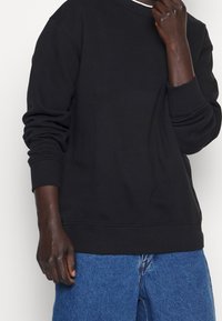 Musta puuvillainen collegepaita, jossa resorireunat hihoissa ja helmassa. Pyöreä kaula-aukko. Päällä vaaleansinisen farkun kanssa, korostaen rentoa istuvuutta.