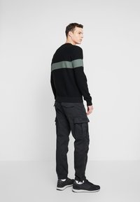 Sort ribbet sweater med en grøn vandret stribe, parret med sorte cargo bukser med flere lommer, suppleret af sorte sneakers.