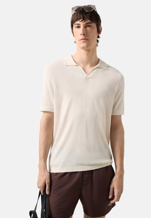 Jonge man met een crème korte mouwen polo, bruine shorts, zonnebril op het hoofd, die zwarte tasgrepen vasthoudt tegen een effen lichte achtergrond.