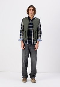 Quilts zaļš vestīts pār zilu un melnu šaha kreklu, pelēki džinsi un bēši apavi; tam ir divas priekšējās kabatas un pogu aproces.