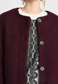 Abrigo burdeos de mezcla de lana con cuello redondo, cierre frontal de botones y detallado encaje negro intrincado sobre un suéter blanco debajo.