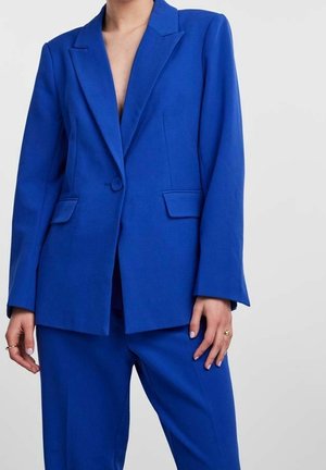 Kobieta nosi jaskrawo niebieski, dopasowany garnitur z marynarką na jeden guzik i dopasowanymi spodniami, stoi na tle gładkiego jasnego tła.