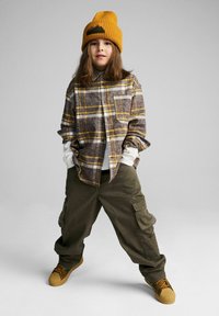 Dziecko ubrane w brązową i żółtą flanelową koszulę w kratę, zielone bojówki i żółtą dzianą czapkę, z brązowymi butami.