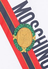 White cotton t-shirt featuring a gold bear emblem enclosed in a green wreath, with bold black and red stripe graphics spelling "MOSCHINO."