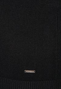 Svart stickat tyg med en texturerad yta, med en liten metallisk etikett med varumärkesnamnet. Designen är enkel och monokrom.