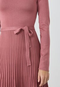 Langermet, ribbestrikket kjole i støvrose med en tettsittende bodice og en plisserte skjørt. Har et stoffbelte knyttet i midjen.