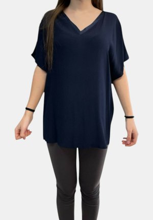 Donna che indossa una tunica blu navy a scollo a V, ampia, con maniche corte e pantaloni attillati scuri, in piedi su uno sfondo semplice.