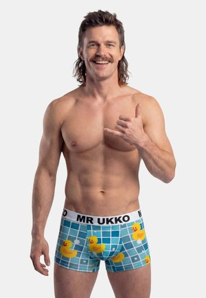 Hombre sonriente sin camisa con mullet y bigote, que lleva calzoncillos tipo bóxer con patrón de patitos de goma, haciendo el gesto de mano shaka.