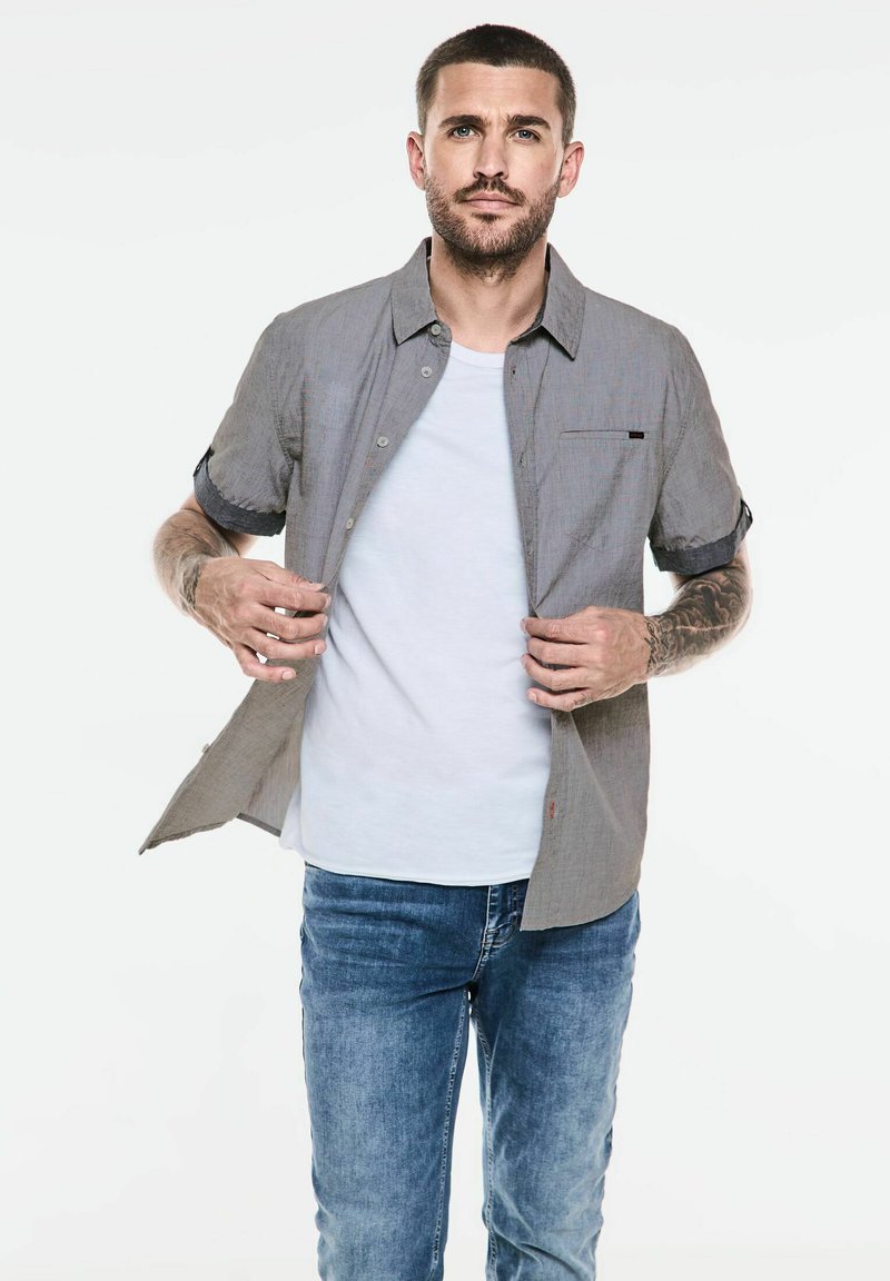 Grå kortärmad skjorta med knappar och bröstficka, bärs över en vit t-shirt och matchas med blå jeans. Slät textur, avslappnad passform.