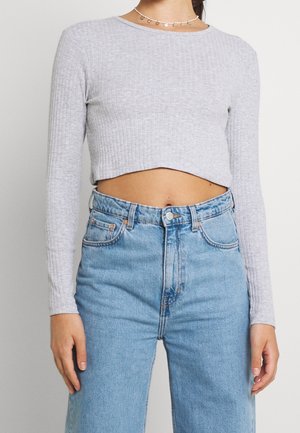 Femme portant un crop top côtelé gris clair à manches longues et un jean taille haute bleu clair, avec un collier en pièce d'or visible autour du cou.