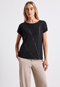 Sort T-shirt med optrukne ærmer, prydet med hvid abstrakt syning, parret med beige bukser. Glat stof, afslappet pasform, enkelt design.