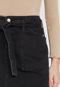 Černá džínová sukně s uvázáním v pase a kapsou na boku, s hladkou texturou a jemným leskem. Zapínání na knoflíky v pase.