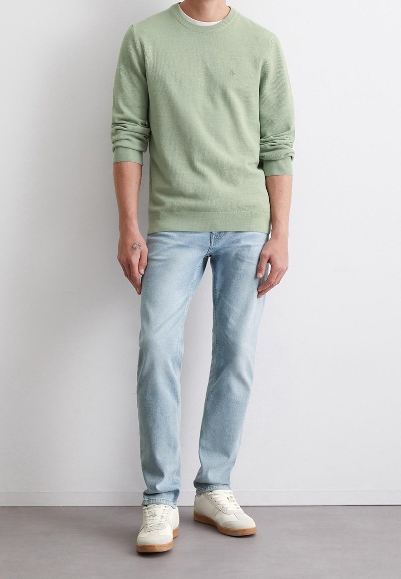 Homme portant un pull vert clair à manches longues, un jean bleu clair et des baskets blanches, debout contre un mur blanc uni.