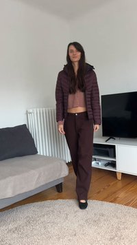 Casaco acolchoado castanho por cima de um suéter rosa curto, combinado com calças largas castanho-escuras e sapatos baixos pretos, num espaço de estar moderno.