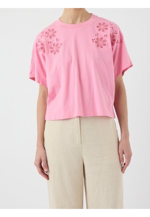 Persoon draagt een losse roze top met korte mouwen met bloemmotieven bij de schouders en lichtbeige broek.