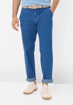 Mand iført opkvaste blå jeans, beige bælte, lyseblå skjorte og hvide sneakers, stående mod en hvid baggrund.