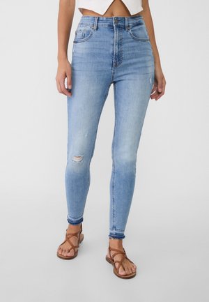 Vrouw draagt lichtblauwe gescheurde skinny jeans en bruine platte sandaaltjes met bandjes op een effen achtergrond.