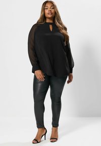 Top nero a maniche lunghe con scollo rotondo e dettaglio a chiave, abbinato a pantaloni neri in similpelle aderenti e sandali neri con tacco.