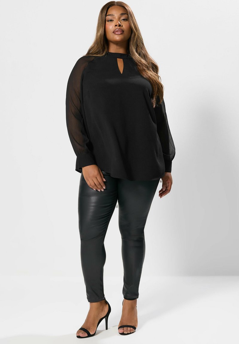 Top nero a maniche lunghe con scollo rotondo e dettaglio a chiave, abbinato a pantaloni neri in similpelle aderenti e sandali neri con tacco.