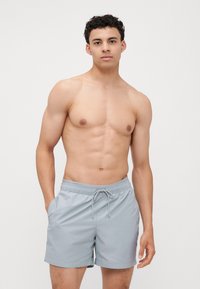 Grijze zwembroek met een elastische tailleband en trekkoord, voorzien van zijzakken en een gladde textuur, gedragen door een model zonder shirt.