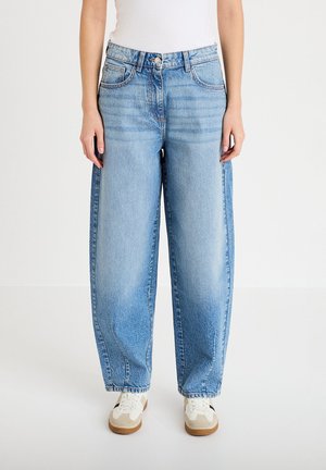 Jasnoniebieskie dżinsy z wysokim stanem i szerokimi nogawkami z przednimi kieszeniami, noszone z białymi trampkami i białym topem, stojące na białej podłodze.