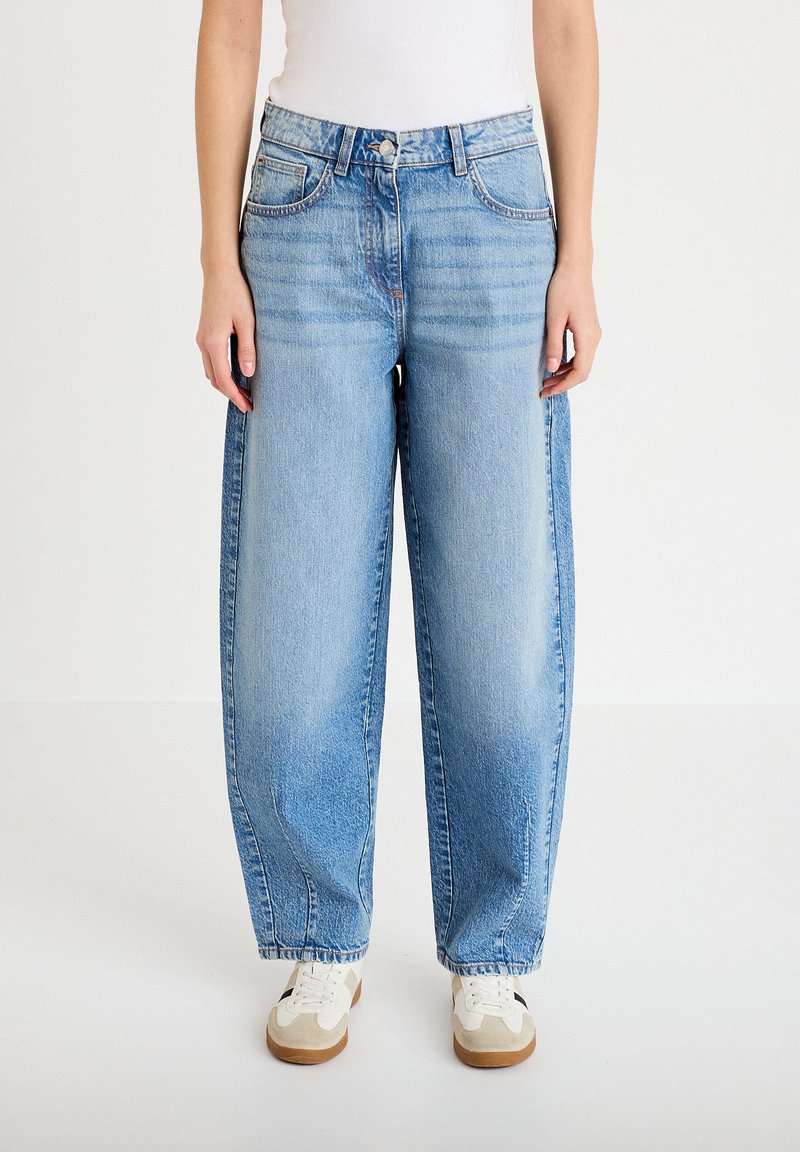 Jeans blu chiaro a vita alta con gambe larghe e tasche frontali, indossati con sneakers bianche e un top bianco, in piedi su un pavimento bianco.