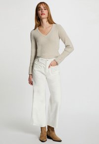 Mujer de pie con la mano en el bolsillo, vistiendo un suéter de punto beige con escote en V, pantalones anchos de talle alto blancos y botas de ante marrones hasta el tobillo.