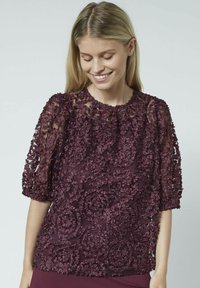 Vinrød bluse med blomsterapplikation, korte puffede ærmer og rund halsudskæring. Transparent blonde tekstur med en flydende pasform.