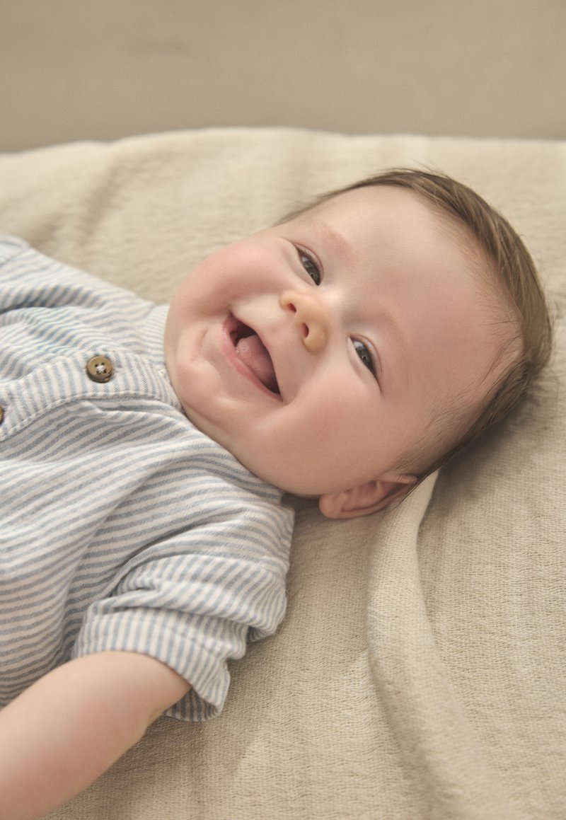 Hellblauer gestreifter Strampler mit braunen Knöpfen an einem lächelnden Baby, das auf einer strukturierten beigen Decke liegt. Sanftes Licht hebt die Gesichtszüge hervor.