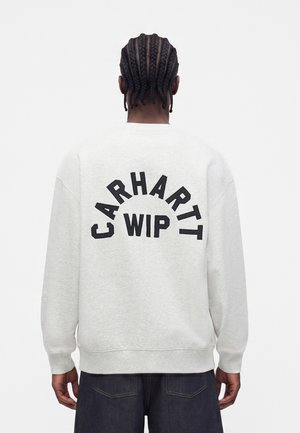 Person med flettet hår set bagfra iført en lysegrå sweatshirt med "CARHARTT WIP" i store sorte bogstaver på ryggen.