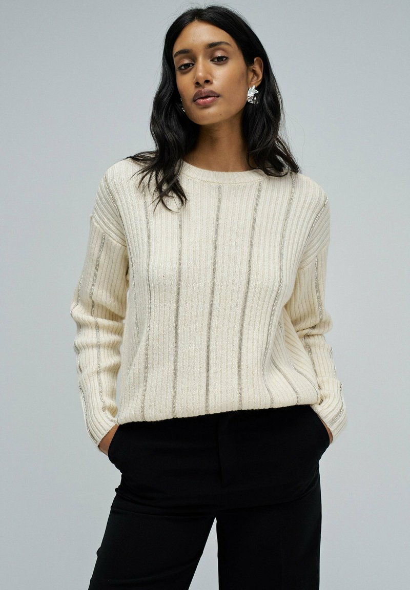 Crème kleurige geribde gebreide trui met zilveren verticale strepen, ronde halslijn en lange mouwen, gecombineerd met zwarte broek. Gestructureerde stof.