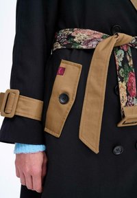 Czarny płaszcz trencz z brązowym paskiem w talii z kwiatowym wzorem, zapinanymi kieszeniami i kontrastującym jasnoniebieskim dzianinowym mankietem.