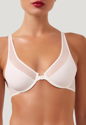 Donna che indossa un reggiseno bianco con chiusura frontale e pannelli in rete trasparente sulle spalle, mostrando la parte superiore del torso e rossetto rosso su sfondo neutro.