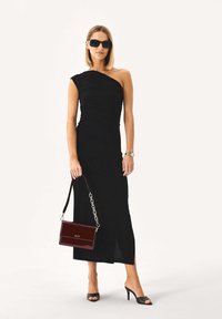 Czarna sukienka na jedno ramię z marszczonym materiałem, z rozcięciem do połowy uda. Modelka trzyma bordową lakierowaną torebkę na łańcuszku i nosi czarne okulary przeciwsłoneczne.