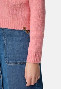 Różowy dziergany sweter z prążkowanymi mankietami, w połączeniu z dżinsami z naszywanymi kieszeniami. Szczegóły obejmują metki z logo na ubraniu.