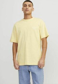 T-shirt di cotone giallo con maniche corte, collo rotondo e testo bianco sul lato sinistro del petto. Abbinato a jeans in denim blu chiaro.