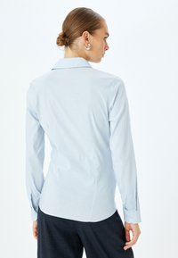Camicia a maniche lunghe azzurro chiaro con colletto a punta, spallina sul retro e vestibilità sartoriale; presenta polsini con bottoni e una texture liscia.