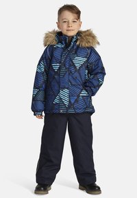 Blå mönstrad jacka med en foder i päls, med geometriska mönster och ränder; matchad med mörka byxor och svarta skor.