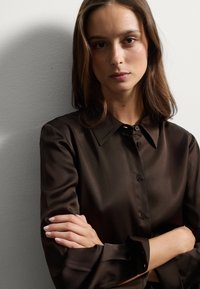 Camisa de satén marrón con botones, cuello y mangas largas, que presenta una textura suave y puños abotonados, llevada con los brazos cruzados.