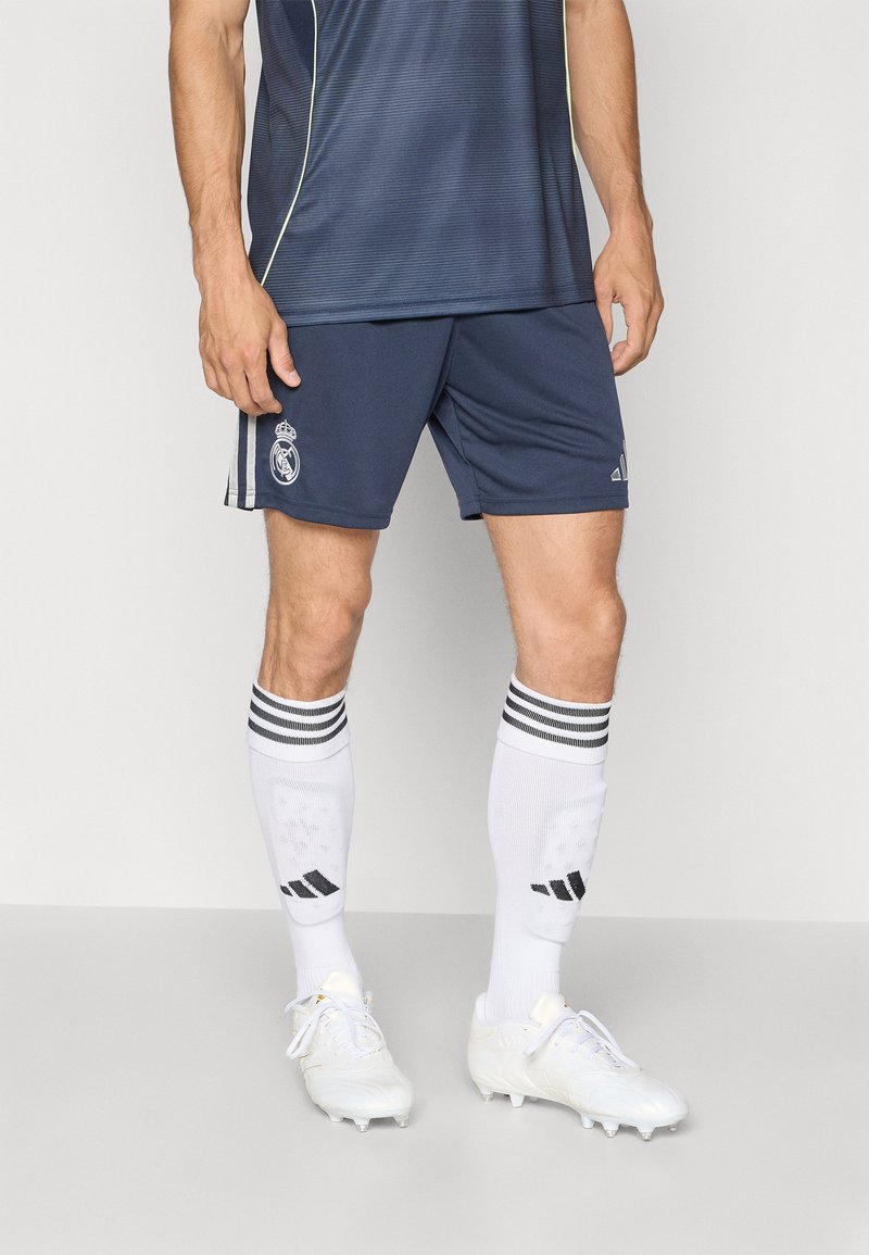 „Adidas Real Madrid“ vyriški futbolo marškiniai, turintys laivyno mėlynumo trumpas rankoves, derančius šortus, baltas kelių kojines su juodais dryžiais ir baltus sportbačius.