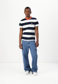 Marineblå og hvitstripet t-skjorte med kort erme og oransje krage; kombinert med løstsittende blå denim cargo bukser og hvite sneakers.