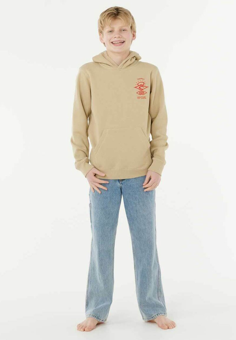 Beige hoodie met een voorvak en rode logo, gecombineerd met lichtblauwe wijde jeans, model staat blootsvoets tegen een effen achtergrond.