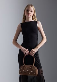 Sac en cuir suédé marron avec des embellissements en forme d'étoile argentée, doté de deux poignées et d'une forme structurée, tenu par un modèle portant une robe noire.