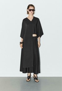 Kvinde iført sort asymmetrisk kjole med udskæring, sorte højhælede sandaler, store solbriller og kraftige armbånd, stående på grå gulv.