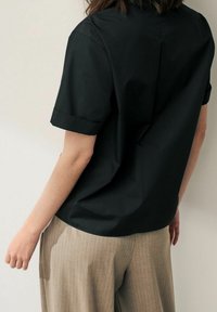 Chemise noire à manches courtes avec une coupe décontractée, en tissu lisse, associée à un pantalon beige clair à fines rayures. Détails de l'ourlet visibles.